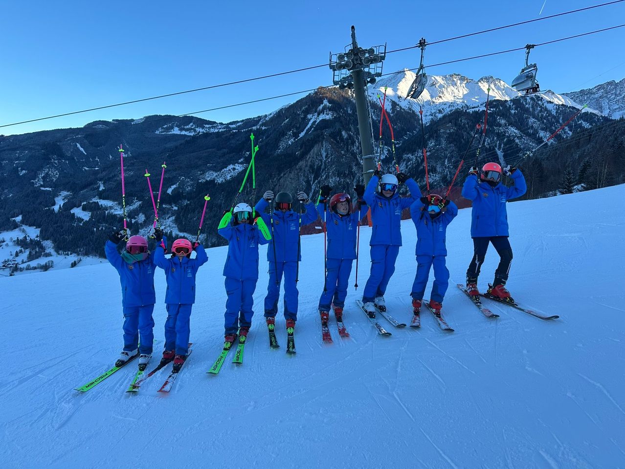 Eine Gruppe von Kindern und Erwachsenen in blauen Skianzügen posiert für ein Foto auf einem verschneiten Berg mit Skiliften im Hintergrund.