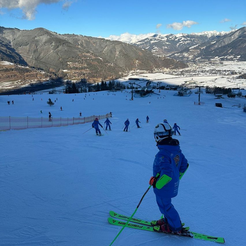 Eine Gruppe von Kindern in blauen Skianzügen rutscht auf einem verschneiten Hang unter blauem Himmel. In der Ferne schneebedeckte Berge.