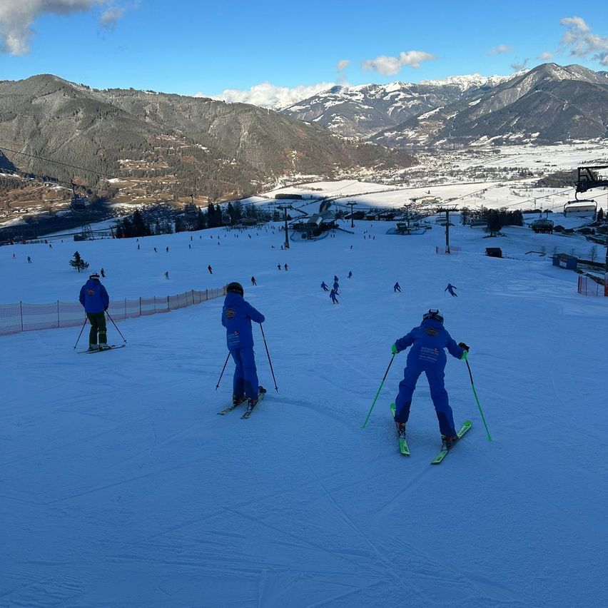 Eine Gruppe von Skifahrern in blauen Skianzügen befindet sich auf einem verschneiten Hang mit Bergen im Hintergrund.