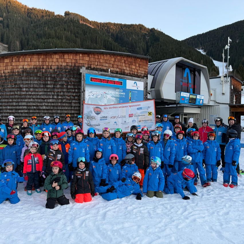 Eine Gruppe von Kindern und Erwachsenen in blauen Skianzügen und Helmen posiert für ein Foto vor einem Ski-Lodge mit Bergen im Hintergrund.