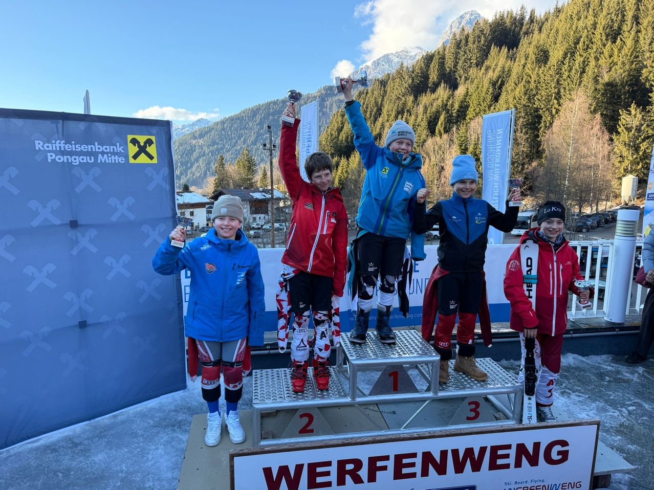 Five children stand on a podium, holding trophies, with snowy mountains in the background. They are dressed in winter gear and appear to be celebrating a victory.