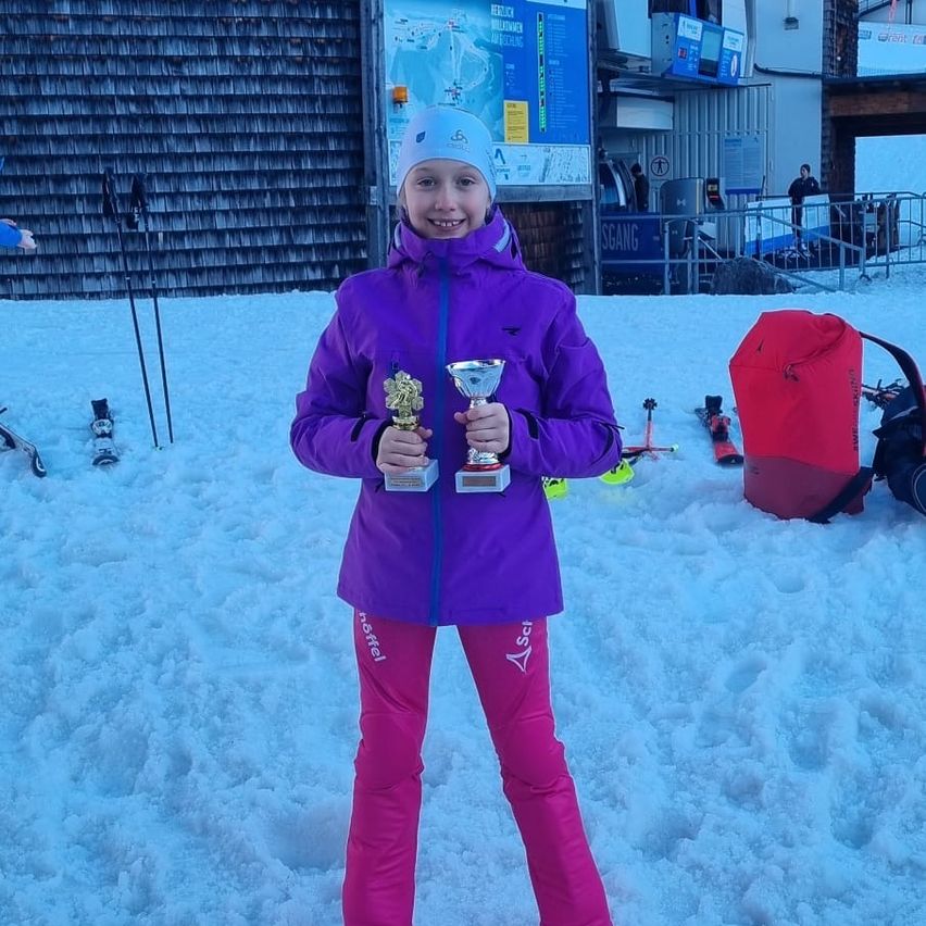 A young girl in a purple jacket poses in the snow, holding two trophies. Skis and a red bag are nearby. In the background, a building with a sign and a person.