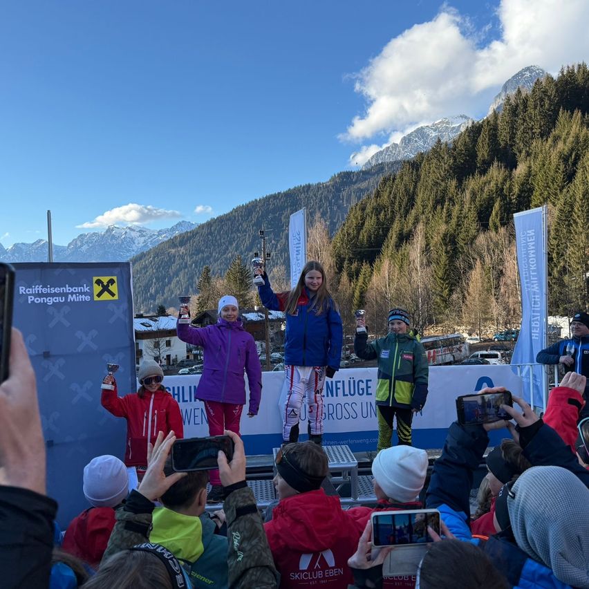 Three young girls stand on a podium, holding trophies. They are dressed in winter sports gear, suggesting they are ski racers. Behind them, a banner reads 'Raiffeisenbank Pongau Mitte'. A mountainous landscape and a blue sky are visible in the background.