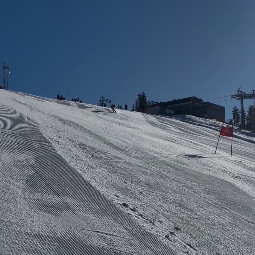 Snowy hill with a few skiers, a red flag, and a ski lift station. The scene is under a clear blue sky.