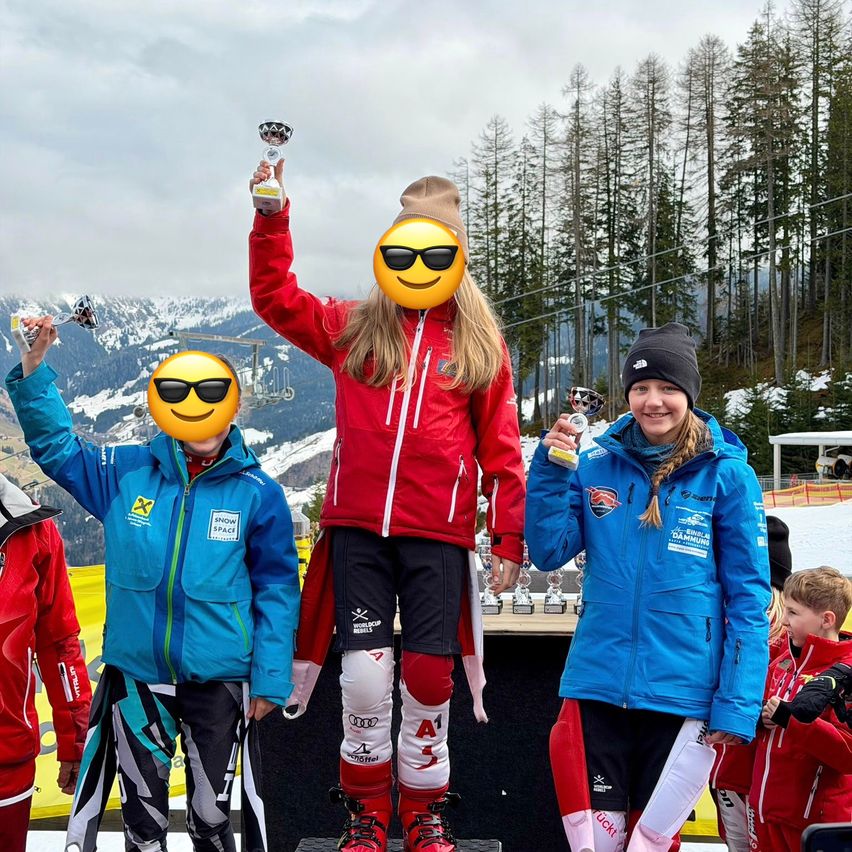 Eine Gruppe von Skifahrern mit Jacken mit Logos steht auf einem Podest und hält Pokale. Ein Skifahrer trägt eine rote Jacke mit einem Smiley-Gesicht-Emoji auf dem Kopf. Verschneite Berge sind im Hintergrund.