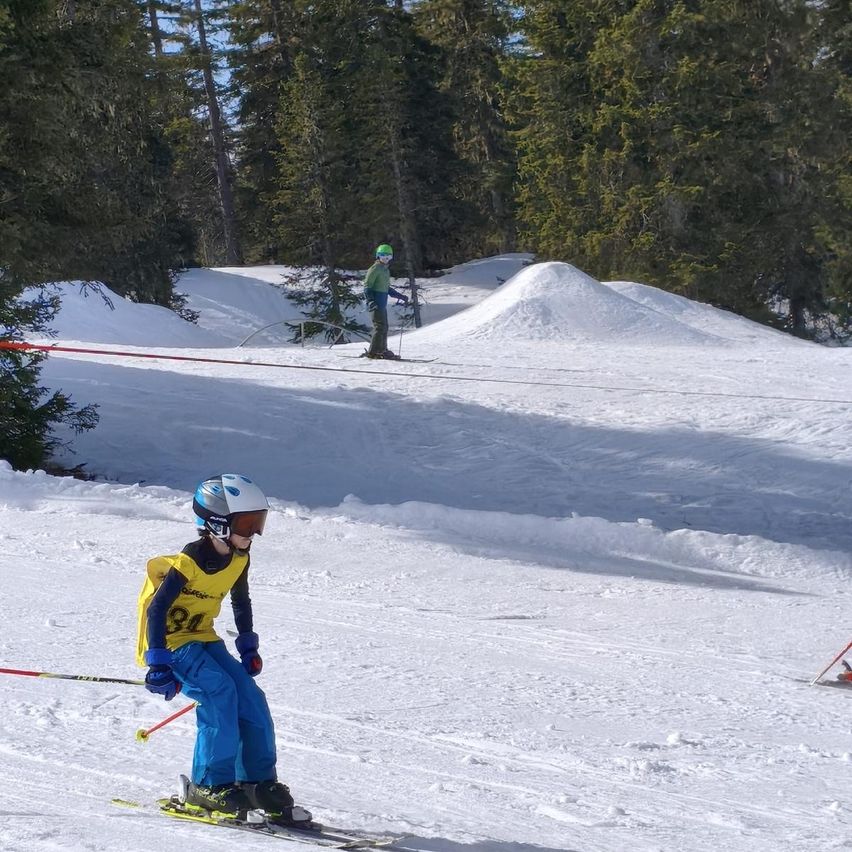 Ein junger Skifahrer, Nummer 34, fährt einen verschneiten Hang hinunter. Dahinter steht eine Person in einer grünen Jacke und Helm in der Nähe einer roten Barriere. Bäume umgeben das Gebiet.