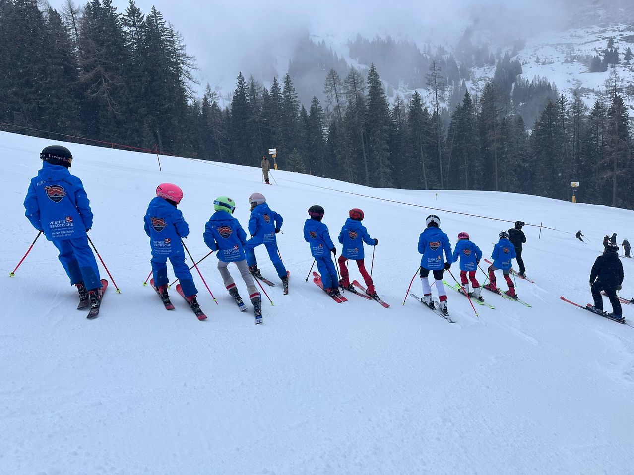 Eine Gruppe von Kindern in blauen Skianzügen mit Helmen und Skistöcken fährt einen schneebedeckten Hang hinunter. Bäume und eine Person sind im Hintergrund.