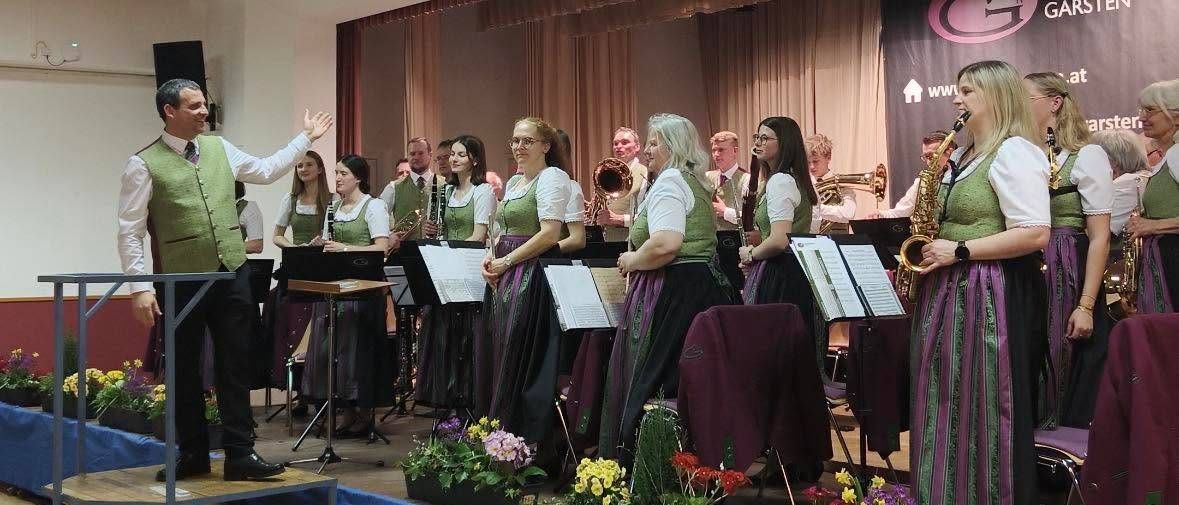 Eine Band tritt auf der Bühne mit verschiedenen Instrumenten auf, gekleidet in traditioneller grüner Kleidung. Sie werden von einem Dirigenten begleitet, und die Bühne ist mit Blumen geschmückt.