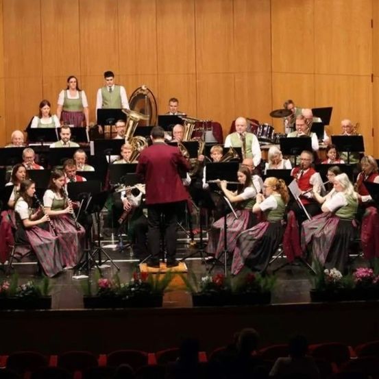 Ein großes Orchester spielt auf der Bühne mit einer Mischung aus Blech-, Holz- und Percussionsinstrumenten. Die Musiker sind in traditioneller Kleidung gekleidet und das Publikum schaut von der ersten Reihe zu.