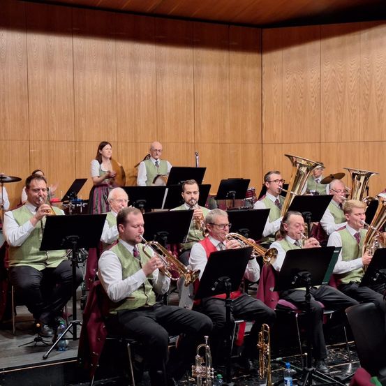 Eine Gruppe Musiker spielt auf der Bühne und musiziert mit Trompeten und Posaunen, mit einer Frau in einem Kleid im Hintergrund.