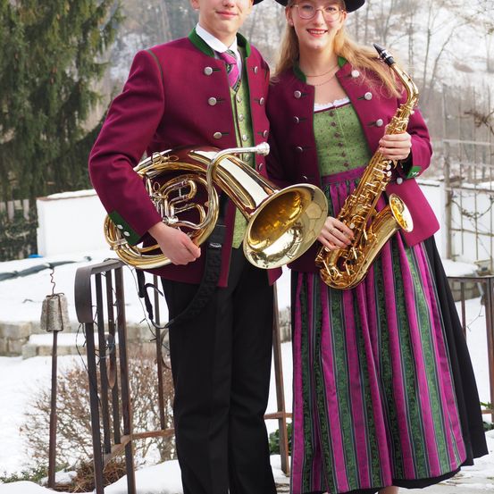 Zwei Menschen in traditioneller Kleidung posieren mit ihren Instrumenten, einer hält ein Horn und die andere ein Saxophon, in einer verschneiten Umgebung im Freien.