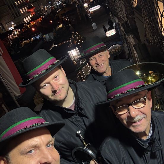 Drei Männer mit schwarzen Hüten und Trompeten posieren für ein Foto in einer festlichen Straßenszene. Sie lächeln und scheinen Musiker zu sein. Der Hintergrund zeigt eine belebte Straße mit festlichen Lichtern und Schildern.