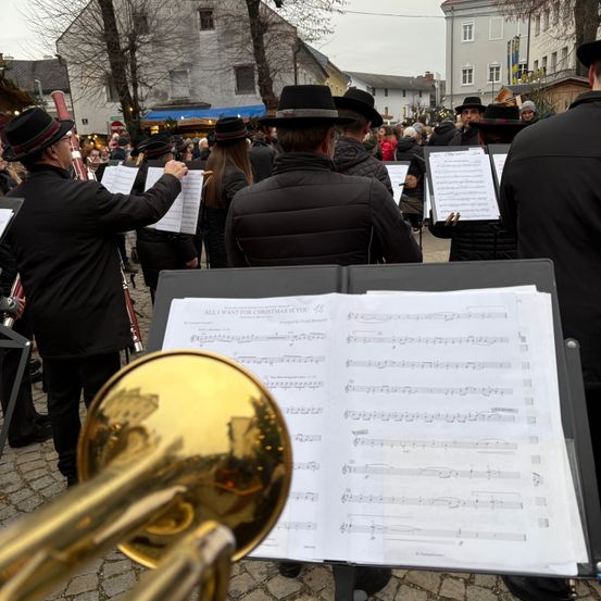 Eine Gruppe von Musikern in schwarzen Hüten und Mänteln tritt in einer Außenumgebung auf, mit sichtbaren Notenblättern und Instrumenten. Eine goldene Trompete ruht auf dem Notenständer.