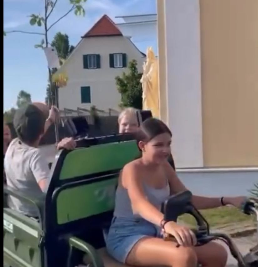Eine junge Frau fährt einen grünen Wagen vor einem Haus mit einer Statue, während andere zusehen und eine Person ein Telefon hält.