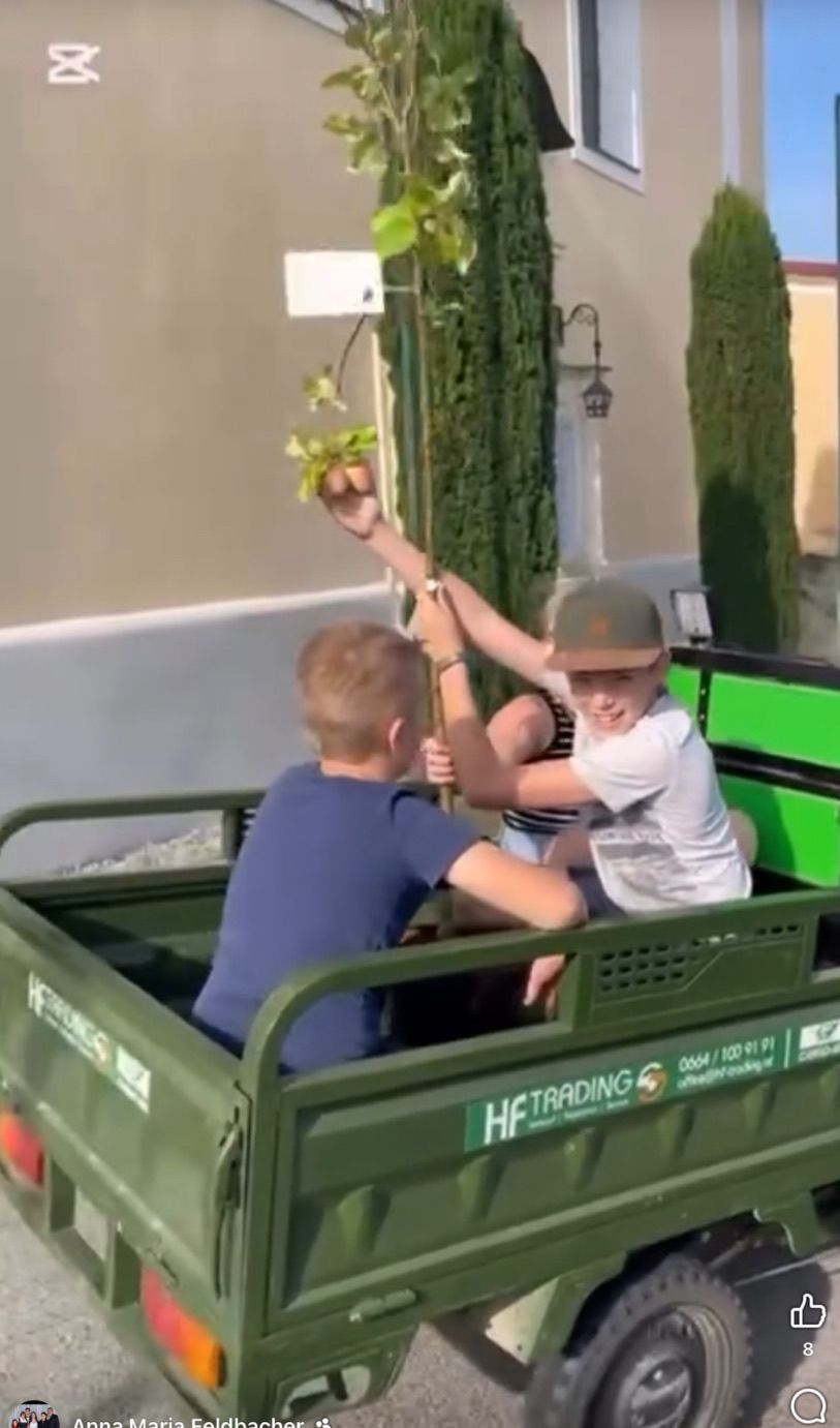 Zwei Kinder sitzen in einem grünen Fahrzeug, eines hält eine Pflanze. Das Kind auf der rechten Seite lächelt. Dahinter steht ein Gebäude mit einer Lampe und einem Baum.