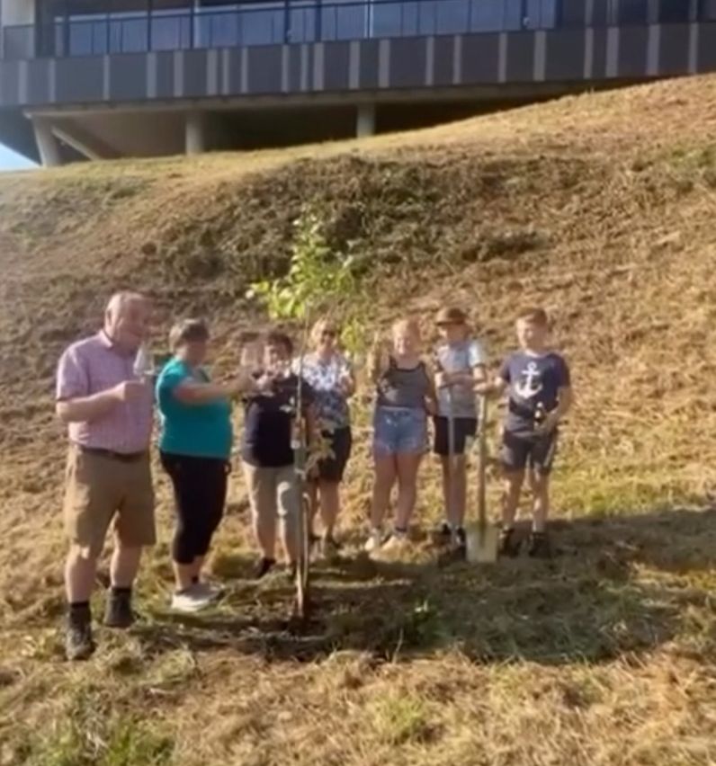 Eine Gruppe von Menschen steht um einen neu gepflanzten Baum und hält Schaufeln und Erde. Ein Gebäude mit großen Fenstern ist im Hintergrund zu sehen.