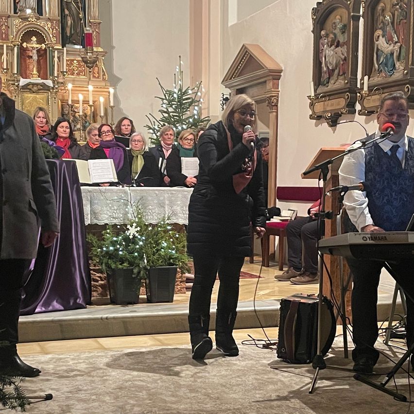 Eine Frau singt in einer Kirche, begleitet von einem Pianisten. Topfpflanzen und ein Weihnachtsbaum schmücken die Bühne. Hinter ihr steht eine Gruppe von Menschen, einige halten Notenblätter in den Händen. Vor dem Pianisten steht ein Mikrofon, neben ihm ein Yamaha-Keyboard.