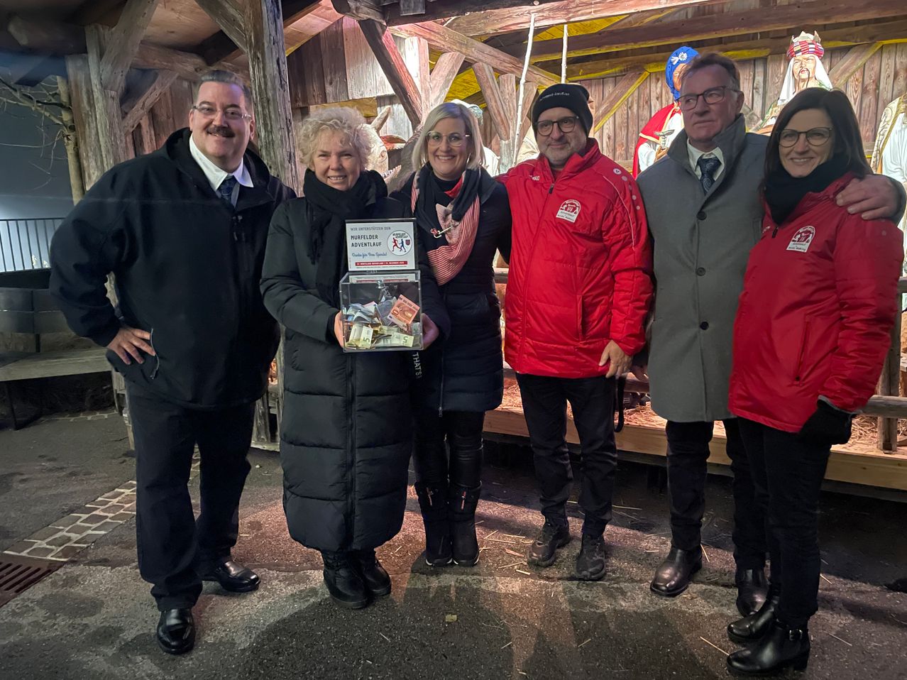 Fünf Menschen posieren für ein Foto. Zwei Frauen in der Mitte halten eine Box mit Geld, und ein Mann in einem roten Mantel hält die Frau am Arm. Der Hintergrund ist eine Holzstruktur.