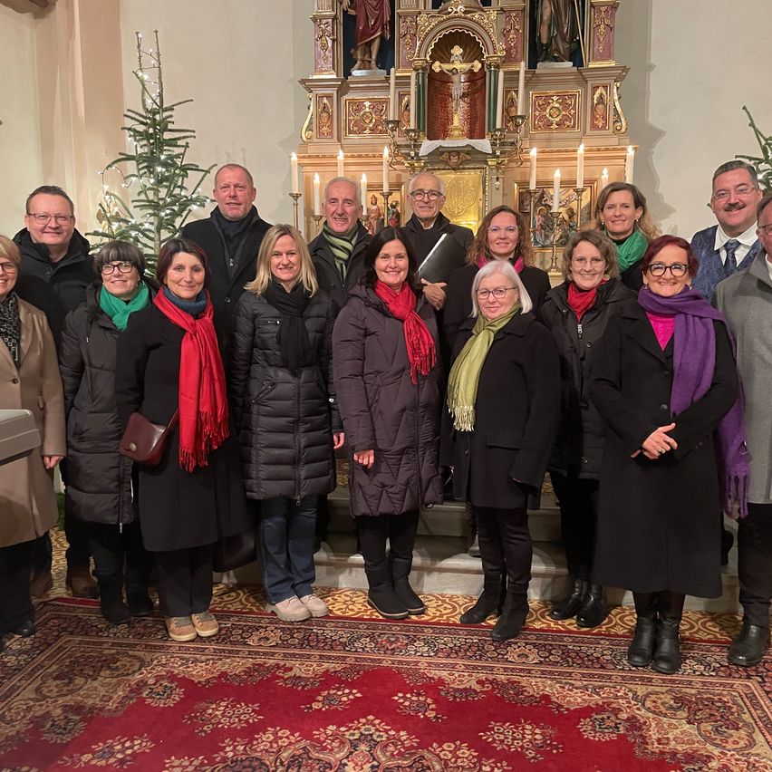 Eine Gruppe von Erwachsenen steht vor einem Altar in einer Kirche und lächelt für ein Foto. Dahinter steht ein Weihnachtsbaum und ein Altar mit Kerzen und religiösen Dekorationen.