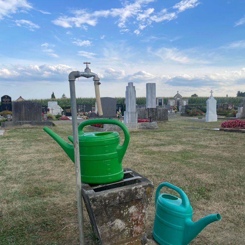 A green watering can is placed on a concrete block with a faucet attached, situated in a cemetery with tombstones and a blue sky in the background.
