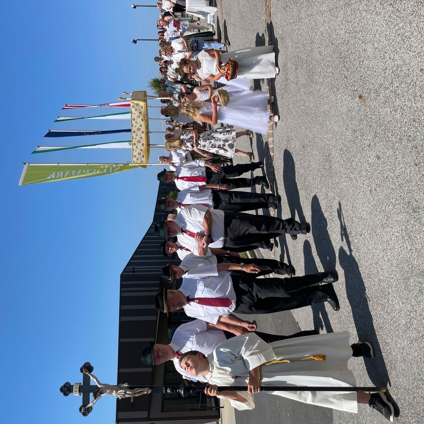 A procession with a priest and choir members lined up under a blue sky. The crowd is dressed in white and red, and flags are displayed in the background.