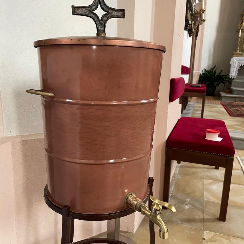 Ein großer kupferner Wasserspender mit einem Kreuz oben, gelegen in einem Kirchenflur. Rote Kissen und eine Pflanze sind im Hintergrund sichtbar.