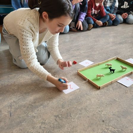 Ein Mädchen hockt auf dem Boden und spielt ein Spiel mit Miniaturtieren. Andere Kinder sitzen im Hintergrund.