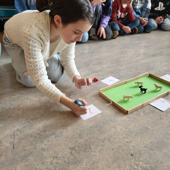 Ein Mädchen hockt auf dem Boden und spielt ein Spiel mit Miniaturtieren. Andere Kinder sitzen im Hintergrund.