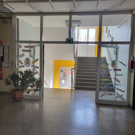 Ein leerer Flur mit zwei Glastüren, die zu Treppen führen. Die Türen haben gelbe und weiße Aufkleber mit Text. Ein Topf mit Pflanzen steht vor den Türen. An der Wand befindet sich ein Feuerlöscher.
