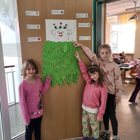 Drei junge Mädchen stehen vor einer Tür mit einem Poster aus grünem Papier. Das Poster hat ein Gesicht und Hände daran befestigt. Sie lächeln.