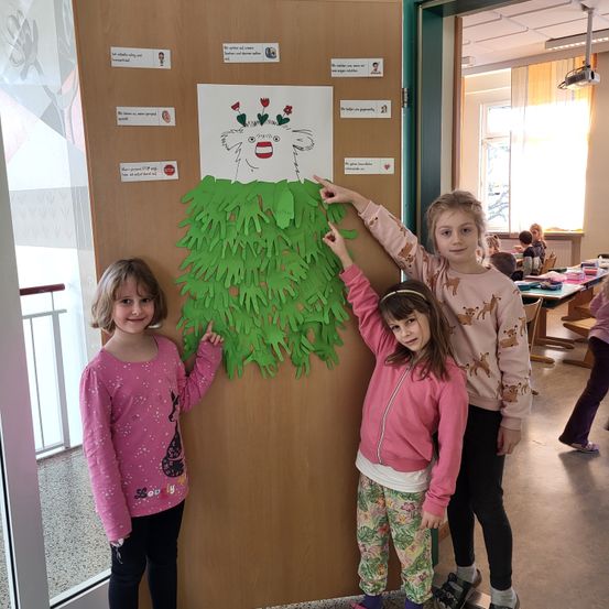 Drei junge Mädchen stehen vor einer Tür mit einem Poster aus grünem Papier. Das Poster hat ein Gesicht und Hände daran befestigt. Sie lächeln.