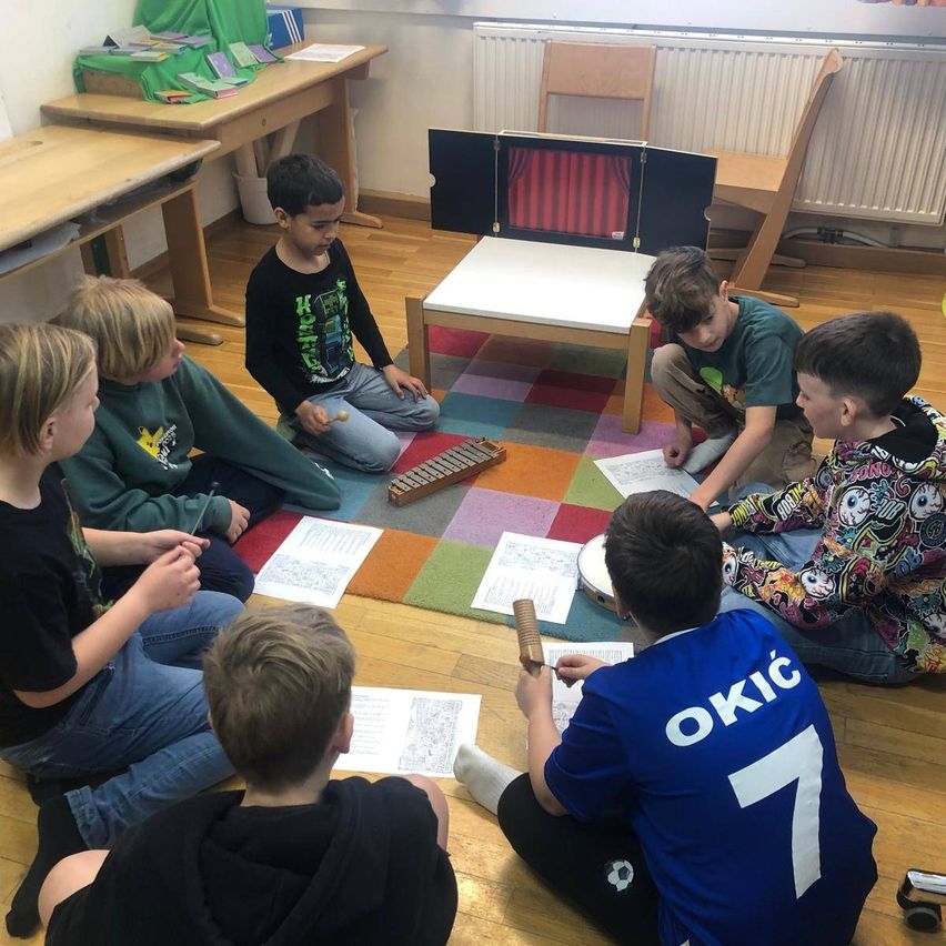 Eine Gruppe von Kindern sitzt im Kreis auf dem Boden, einige spielen Musikinstrumente und andere schauen auf Papiere. Sie sind in einem Klassenzimmer.