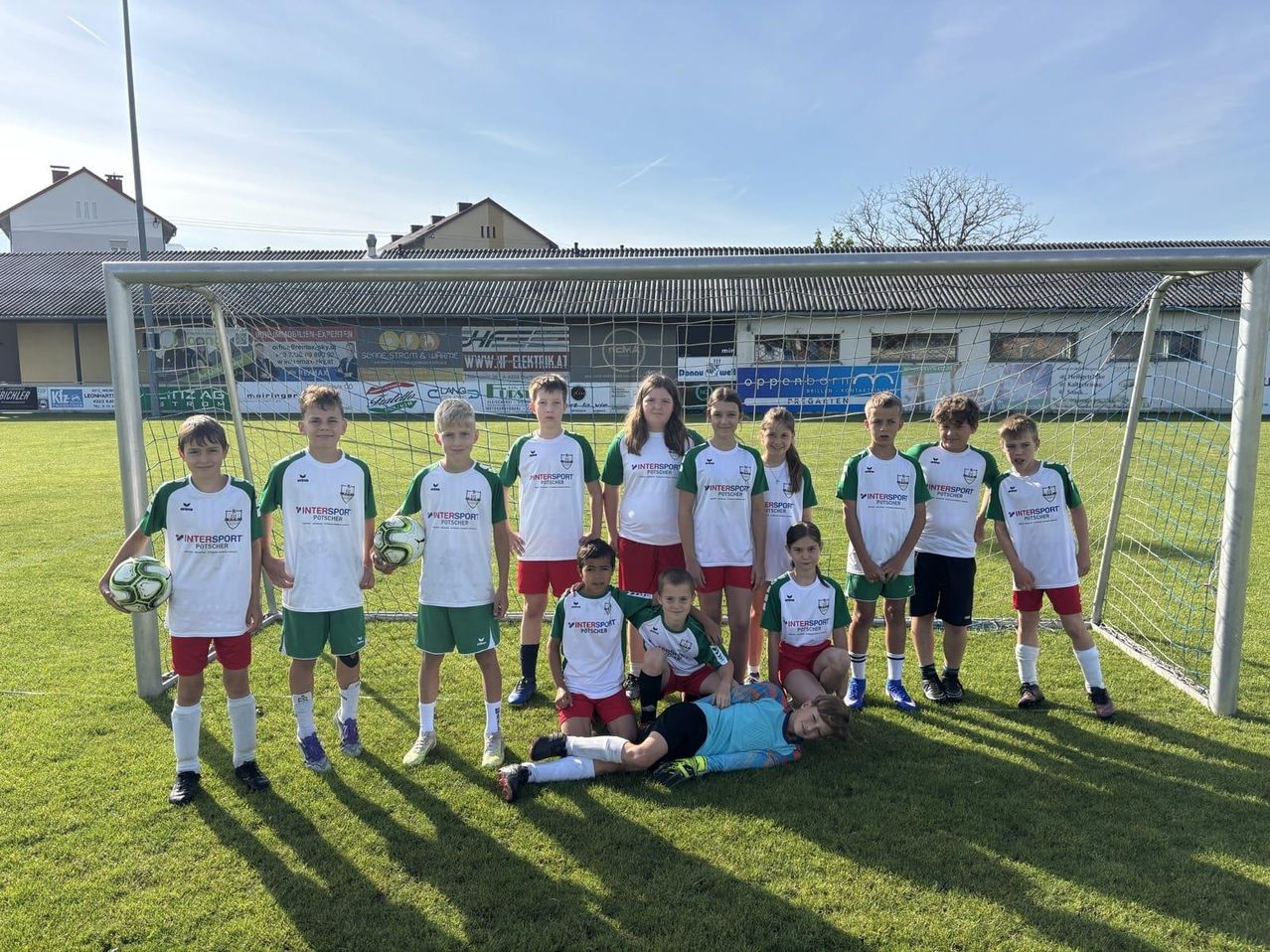Eine Fußballmannschaft mit Spielern in weißen und grünen Trikots steht vor einem Tor. Einige Spieler halten Fußballbälle. Ein Junge liegt auf dem Boden, möglicherweise müde.