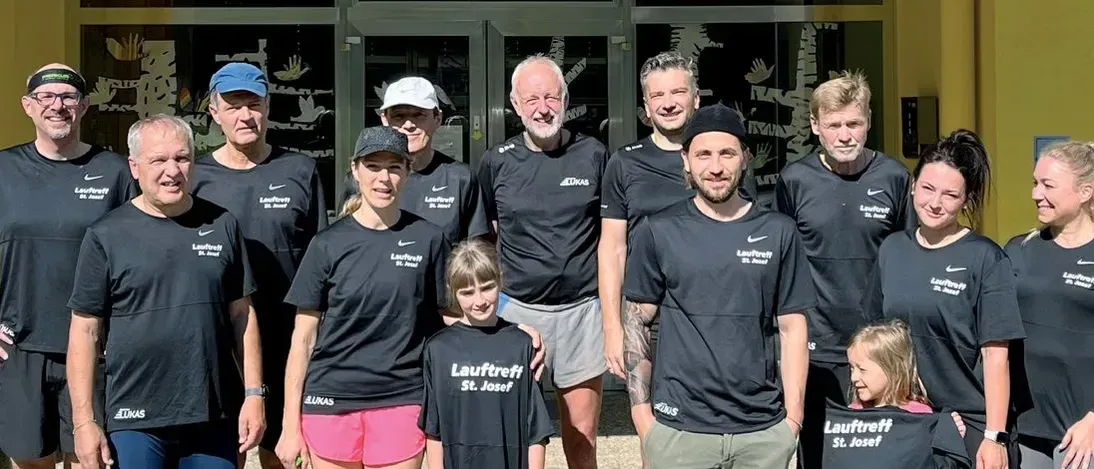 Eine Gruppe von Menschen in passenden schwarzen T-Shirts mit Lauftreff St. Josef darauf lächeln für ein Foto. Sie stehen vor einem Gebäude.