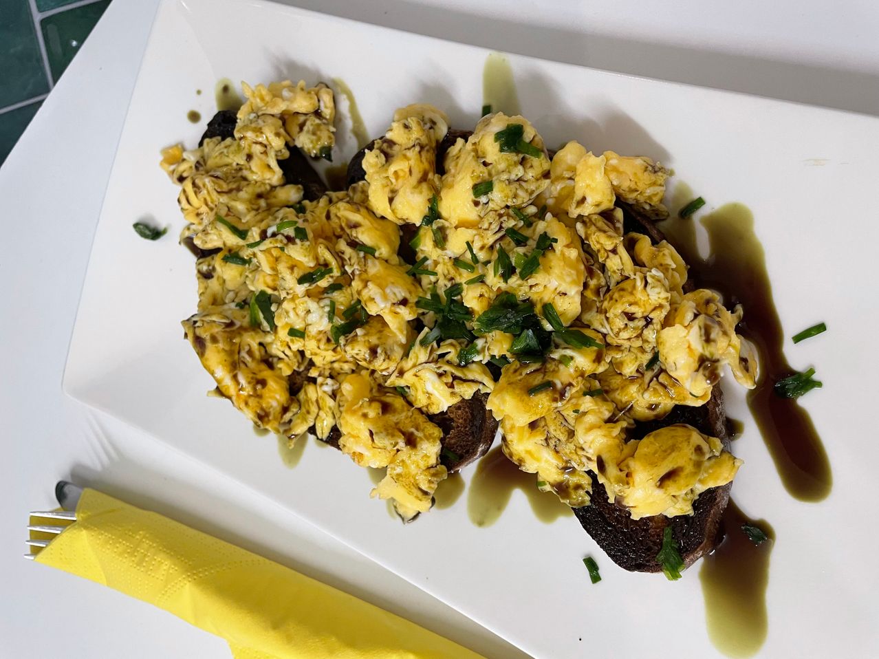 Ein weißer Teller mit Rührei auf Toast, garniert mit grünen Kräutern und einem Saucenüberzug, begleitet von einem gelben Tuch.