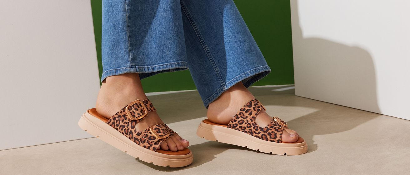 Eine Person in blauen Jeans und Leopardenmuster-Sandalen mit Schnallen und dicker Sohle.