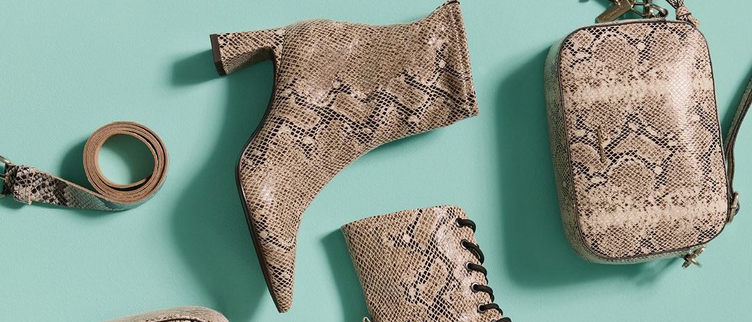 Eine Auswahl an Schuhen, darunter schwarze Stiefel, braune Schlangenmuster-Ankle-Boots, ein Gürtel und eine Umhängetasche auf blauem Hintergrund.