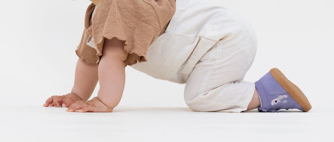Ein Baby mit einem braunen Stirnband und einem braunen Oberteil krabbelt auf einer weißen Oberfläche und trägt weiße Overalls und lila Schuhe mit einem Hasendesign.