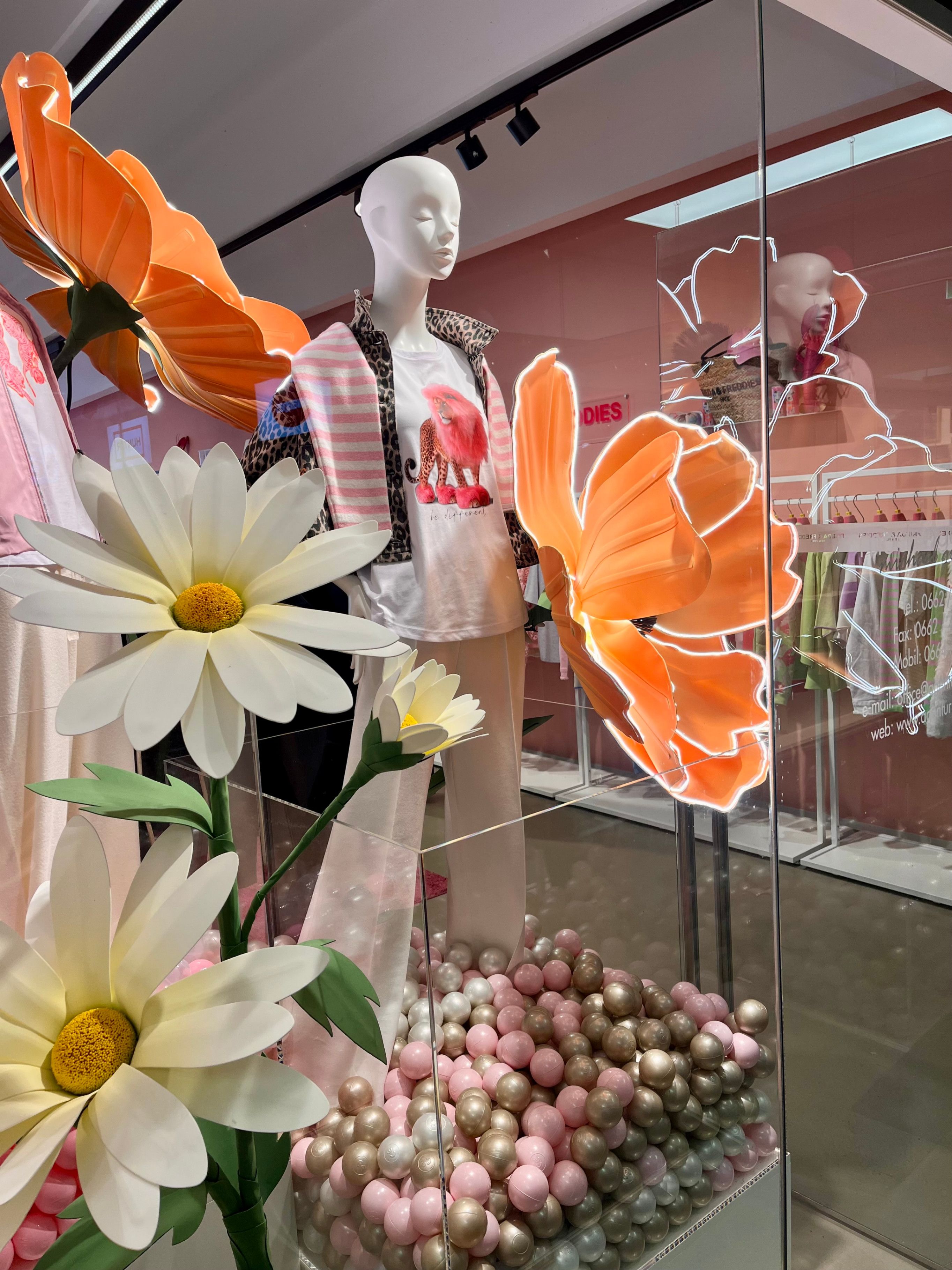 Eine Puppe in einer pink-weiß gestreiften Jacke und einem Elefanten-T-Shirt steht in einem Schaufenster mit künstlichen Blumen und einer großen orangefarbenen Schmetterling.