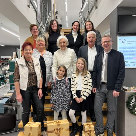 Eine Gruppe von Erwachsenen und Kindern steht auf einer Treppe in einem Geschäft, lächelt und posiert für ein Foto. Sie halten Geschenkboxen, und rechts ist eine Weihnachtsgirlande angebracht.