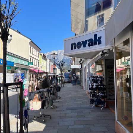 Eine Straße mit Geschäften auf beiden Seiten. Das 'novak'-Geschäft ist prominent mit Taschen und Schuhen im Schaufenster zu sehen. Die Straße ist leer, und ein Lieferwagen ist in der Nähe geparkt.