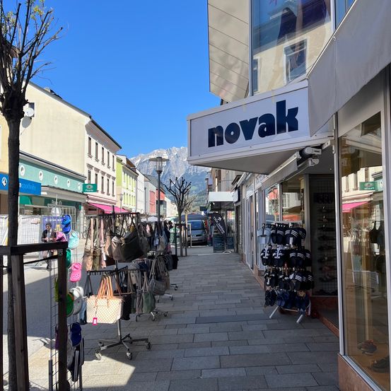 Eine Straße mit Geschäften auf beiden Seiten. Das 'novak'-Geschäft ist prominent mit Taschen und Schuhen im Schaufenster zu sehen. Die Straße ist leer, und ein Lieferwagen ist in der Nähe geparkt.