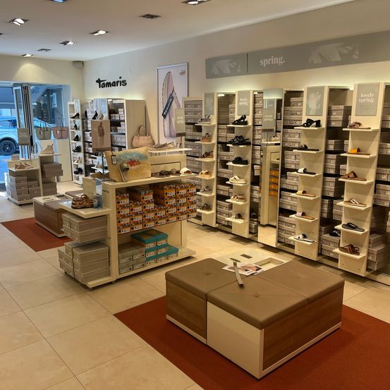 Ein Schuhgeschäft zeigt verschiedene Schuhe auf Regalen, mit einem Schild, das 'Frühling' ankündigt. Schuhe, Handtaschen und Kisten sind ordentlich aufgestellt, und ein Auto ist außerhalb des Eingangs sichtbar.