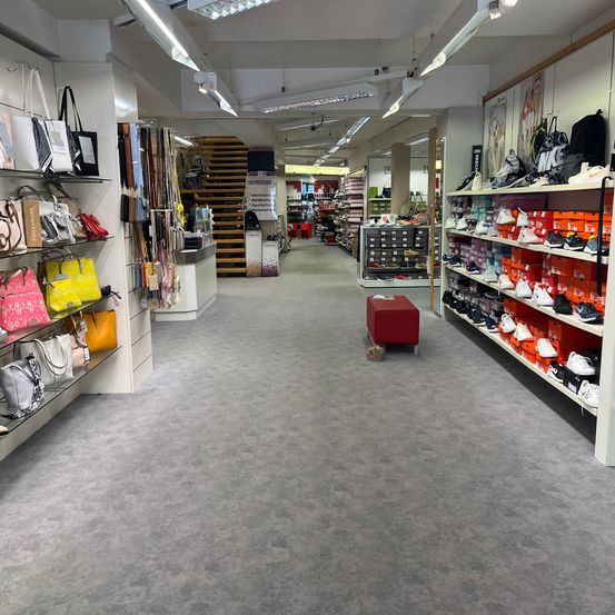 Ein Geschäft mit verschiedenen ausgestellten Artikeln, darunter Handtaschen, Schuhe und Accessoires. Der Laden hat einen grauen Boden und weiße Wände. Die Regale auf der linken Seite haben Handtaschen in verschiedenen Farben und Größen. Die Regale auf der rechten Seite haben Schuhe in verschiedenen Farben und Größen. Die Decke hat mehrere Lampen.