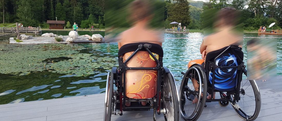 Zwei Personen in Rollstühlen, einer mit orangefarbenem Design und der andere mit blauem, sitzen am See. Hinter ihnen ist eine Holzstruktur und Menschen zu sehen.