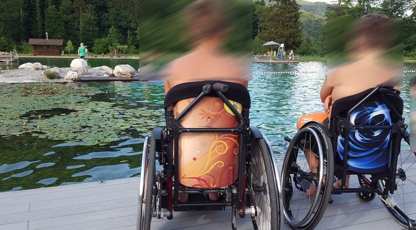 Zwei Personen in Rollstühlen, einer mit orangefarbenem Design und der andere mit blauem, sitzen am See. Hinter ihnen ist eine Holzstruktur und Menschen zu sehen.