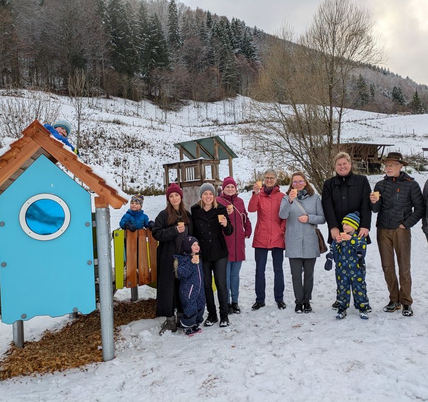 A family with several children, some holding cups, stands in a snowy landscape with a snow-covered hill and forest in the background.