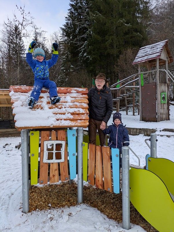 An elderly man and two children stand in front of a wooden playhouse with a snow-covered roof. The children are wearing winter clothes and hats. The man is smiling and holding hands with the child on the right.