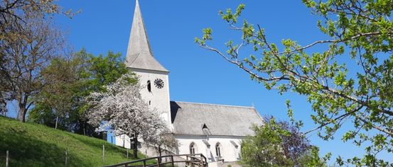 Eine weiße Kirche mit einem Turm und einer Uhr, umgeben von saftigem grünem Gras und Bäumen. Die Kirche steht auf einem Hügel unter einem klaren blauen Himmel.