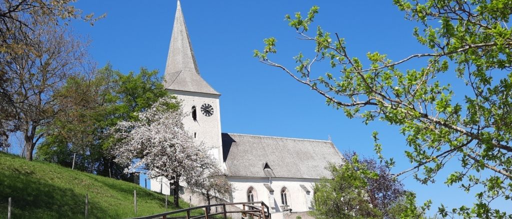Eine weiße Kirche mit einem Turm und einer Uhr, umgeben von saftigem grünem Gras und Bäumen. Die Kirche steht auf einem Hügel unter einem klaren blauen Himmel.